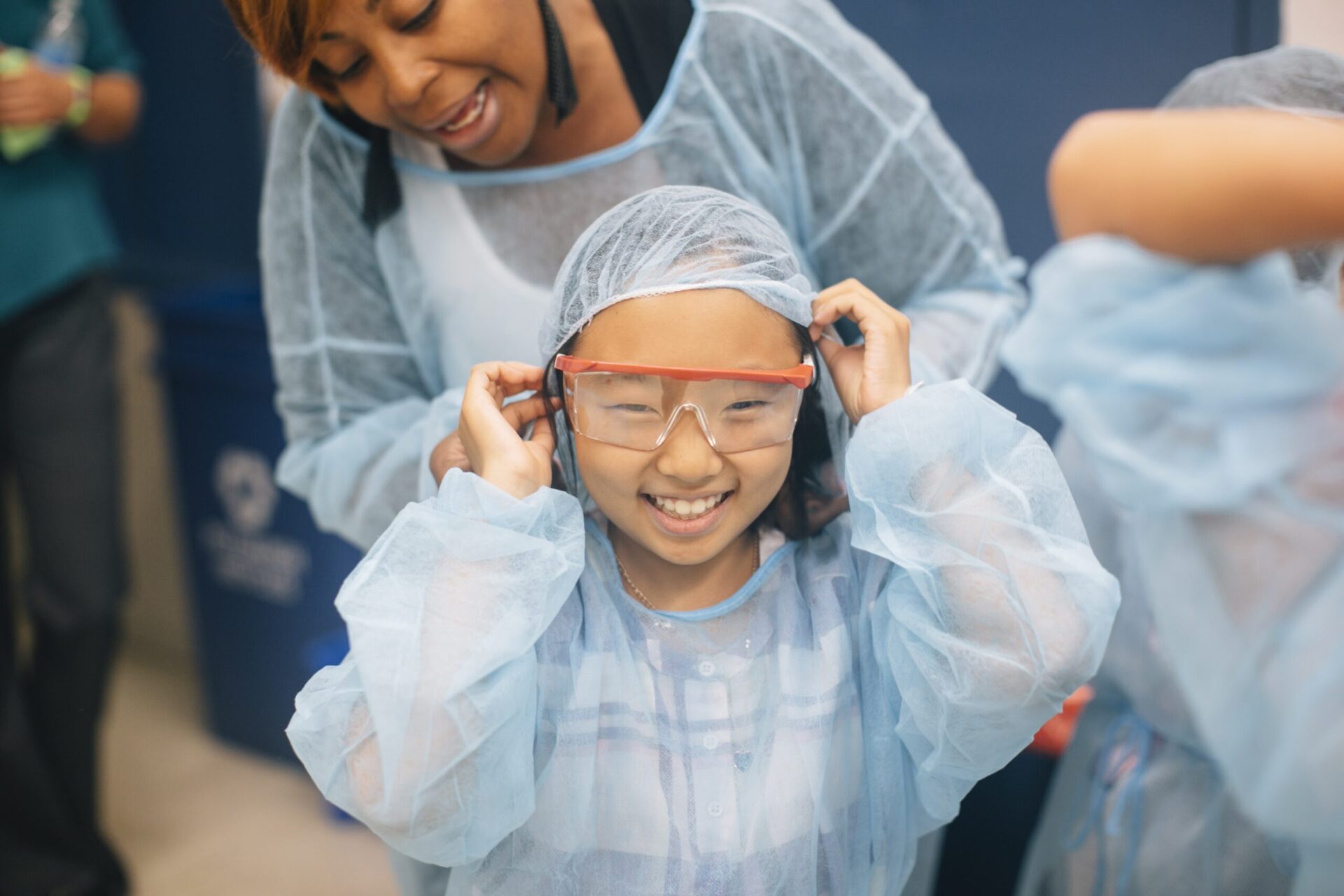 A STEM teacher helps a young student dress in a lab gown during a science lesson.