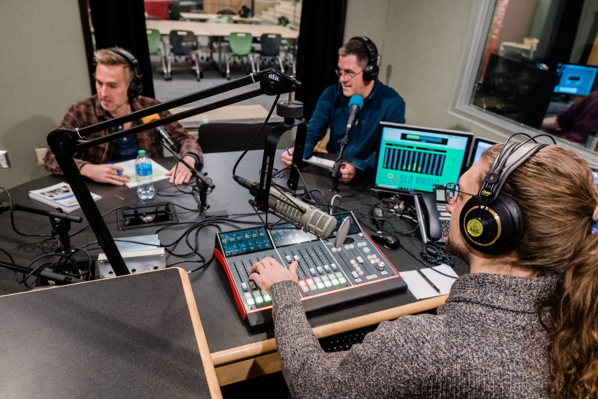 Two men speak into microphones in a recording studio while a third man works on an audio mixing board.