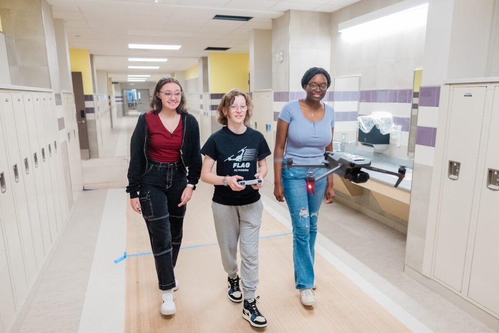 Three teenage students walk down the hall of their school, piloting a drone ahead of them