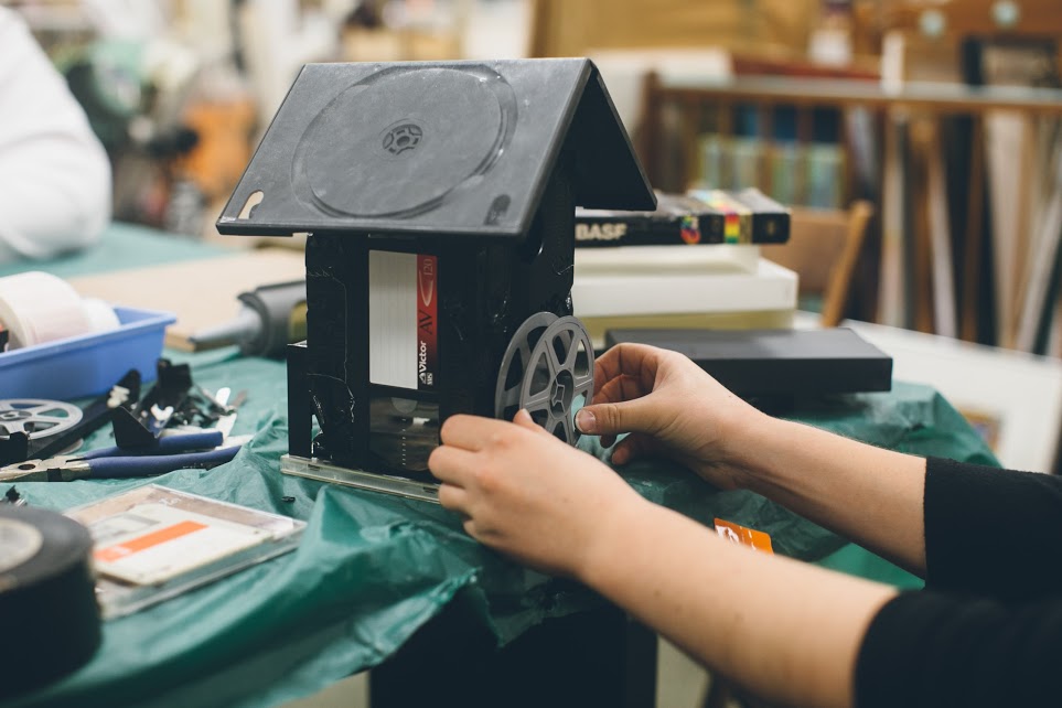 A person assembling a birdhouse from various forms of old media: a dvd case, vhs tapes and a film reel.