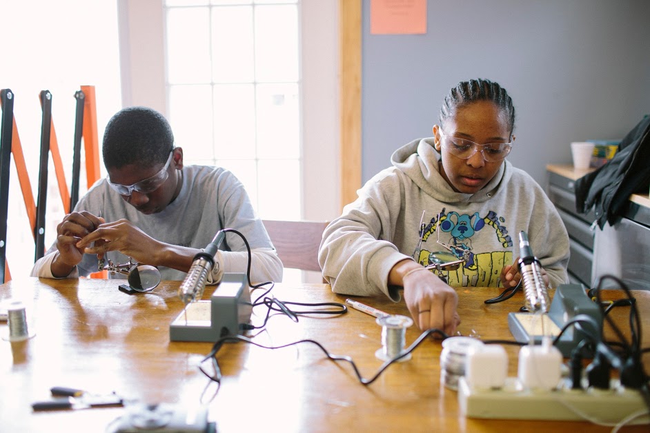 Two teens working at a table with soldering irons.