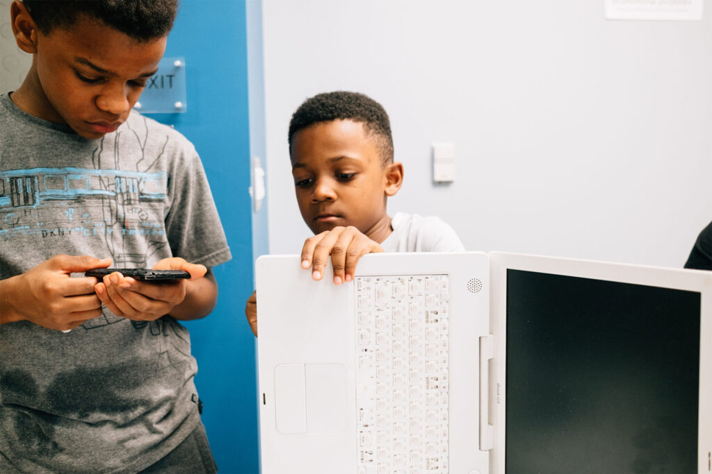 Two young children inspecting a cellphone and a laptop.