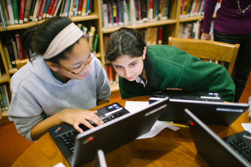 Two girls sit at laptops in a library working on a coding project together