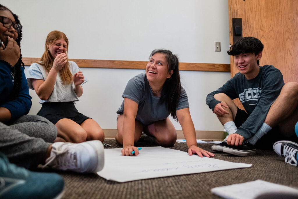A group of teenagers laugh together as they sit on the floor surrounding a large sheet of paper they are writing on.