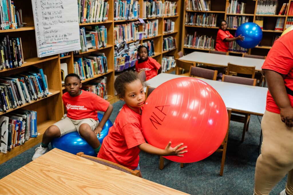 A young girl carries an inflatable exercise ball during a physical therapy learning activity.