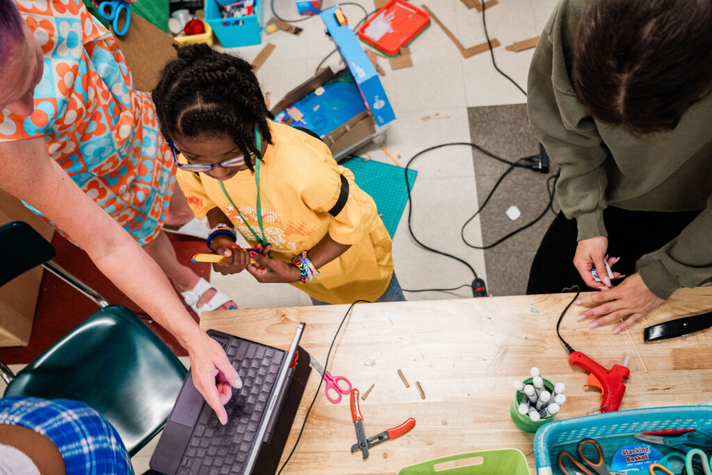 Two middle school students work on a table top to program a pinball machine while an adult educator points the way.