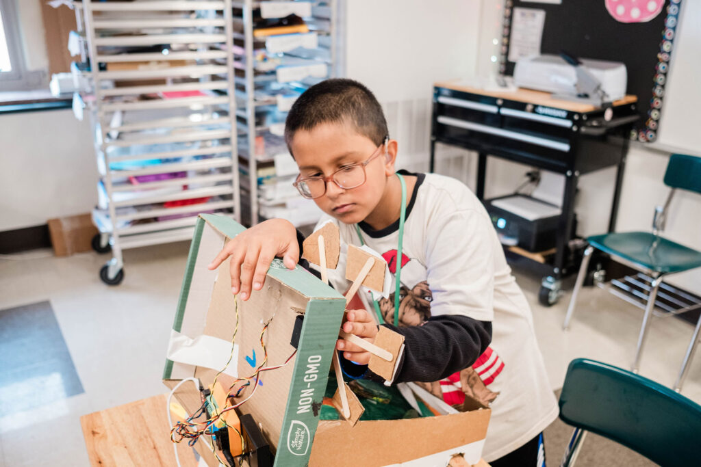 A middle school boy works on the wiring of a do-it-yourself cardboard pinball machine.