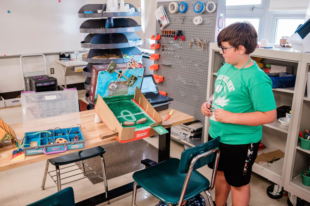 A middle school boy stands beside a pinball machine he built out of cardboard and electronics.