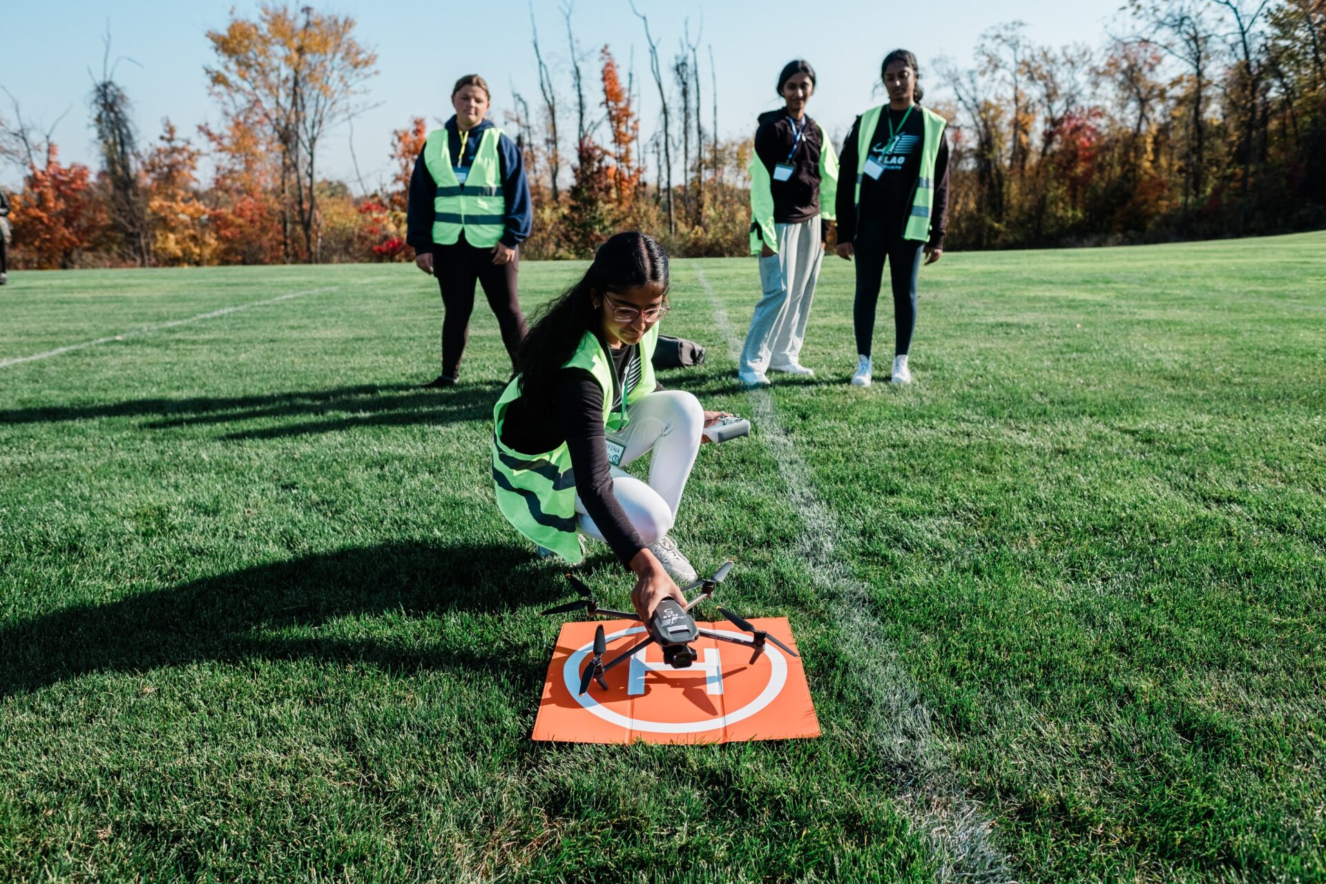 A high school girl kneeling on an outdoor sports field to set up a drone for flight while three other high school girls stand behind her watching.