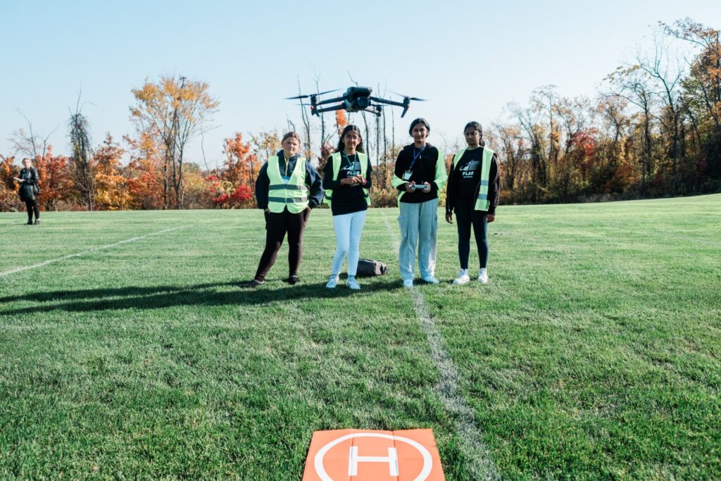 Four high school girls observing a drone aircraft taking off on an outdoor sports field.