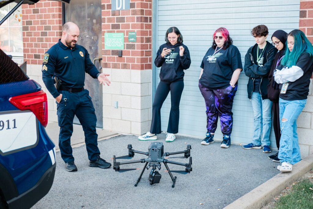 A police officer speaks to a group of high school girls and gestures to a search and rescue drone on the ground.