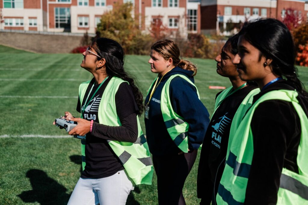 A group of high school girls watching a drone in flight on an outdoor sports field.