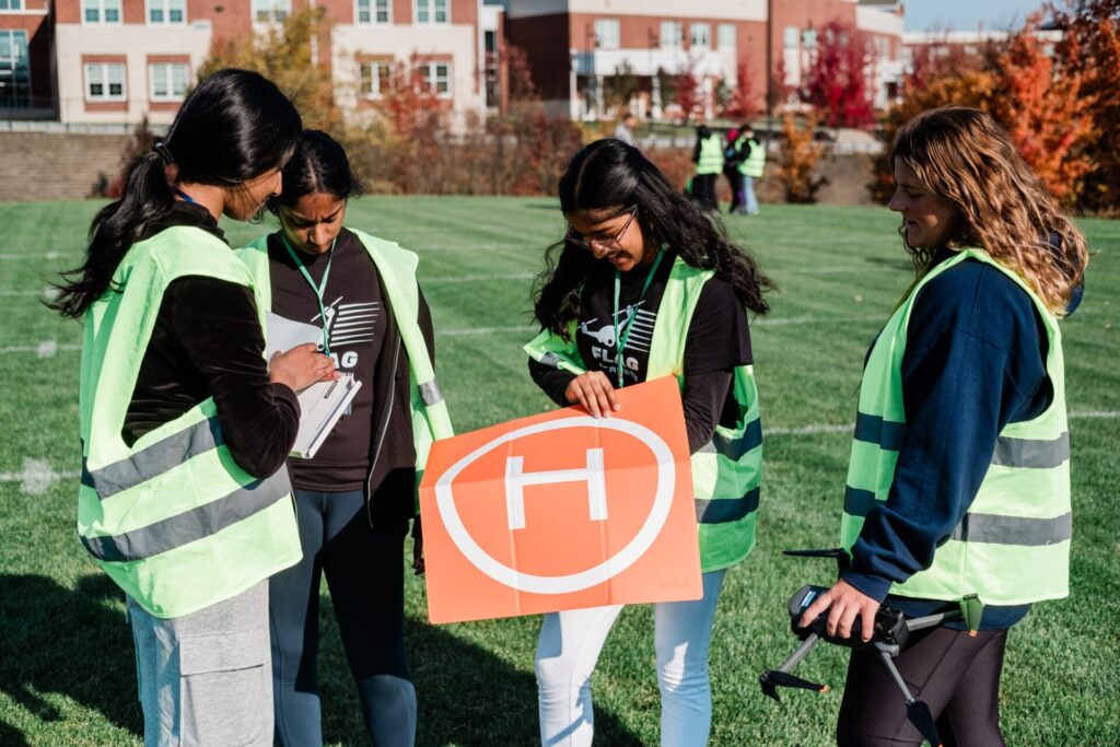 A group of high school girls handling a landing pad for a drone aircraft on an outdoor sports field.