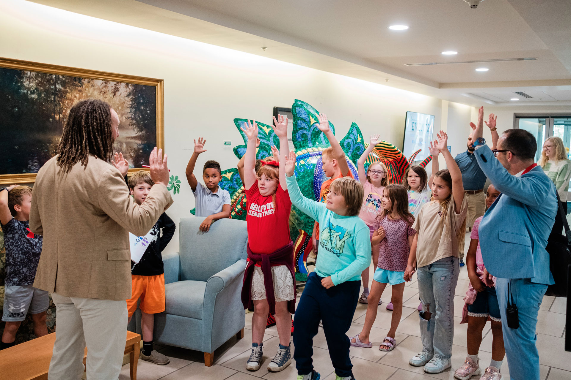 A group of school students assembled in a hospital lobby raise their hands.