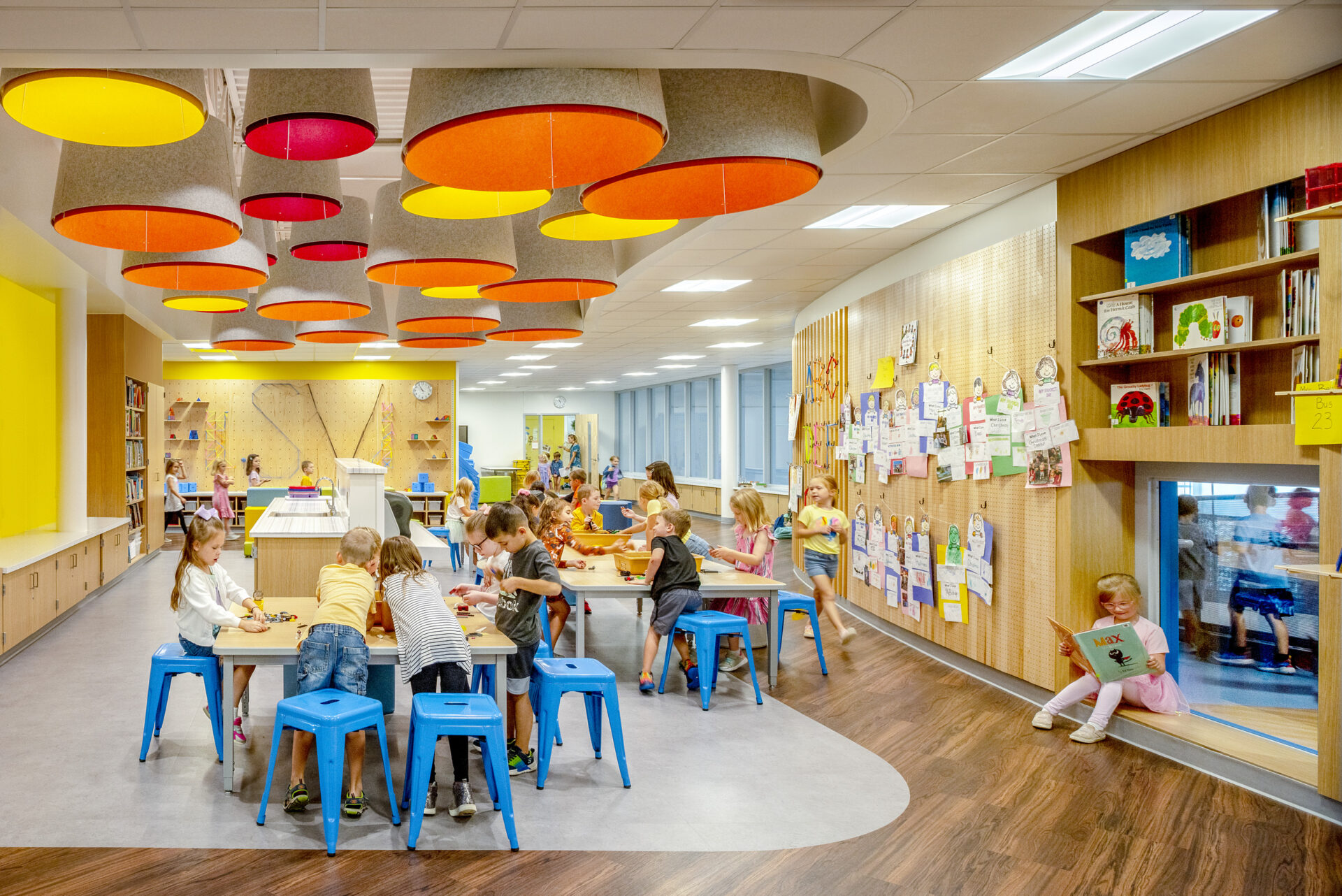 Elementary school aged children sit and stand in small groups around shared work tables inside a bright, colorful school room.