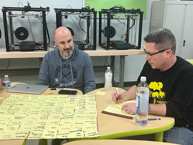 Two men sit at a table and write notes in front of a poster made of sticky-notes.