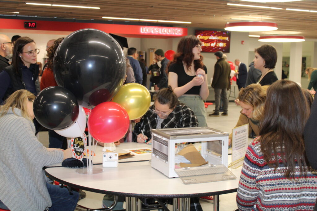A group of high school students crowd around a table decorated with balloons.