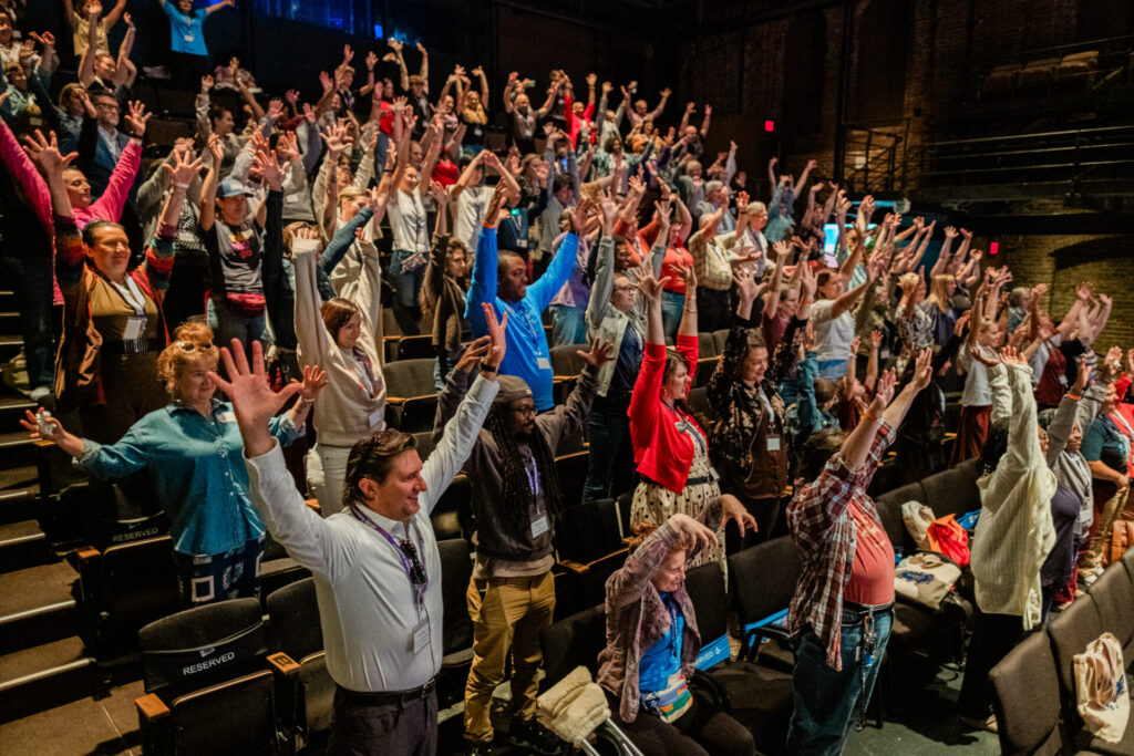 A smiling crowd of people standing in an auditorium with their hands raised above their heads.