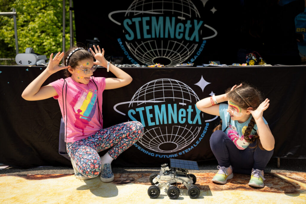 Two girls squat down and hold their hands up like antennae on their head next to a small robot.