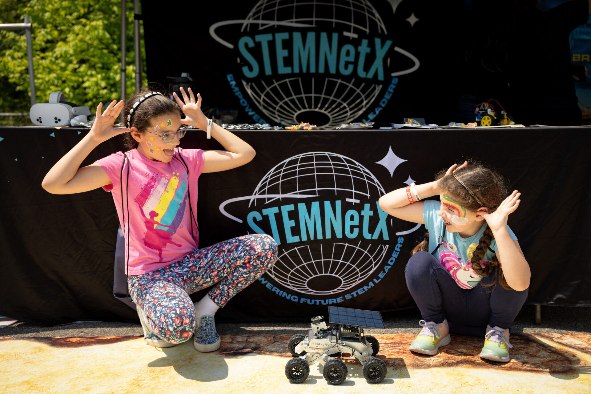 Two girls squat down and hold their hands up like antennae on their head next to a small robot.